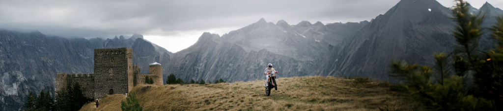 Ein Motorradfahrer fährt auf einem KTM durch eine beeindruckende Berglandschaft mit einer alten Burg im Hintergrund.